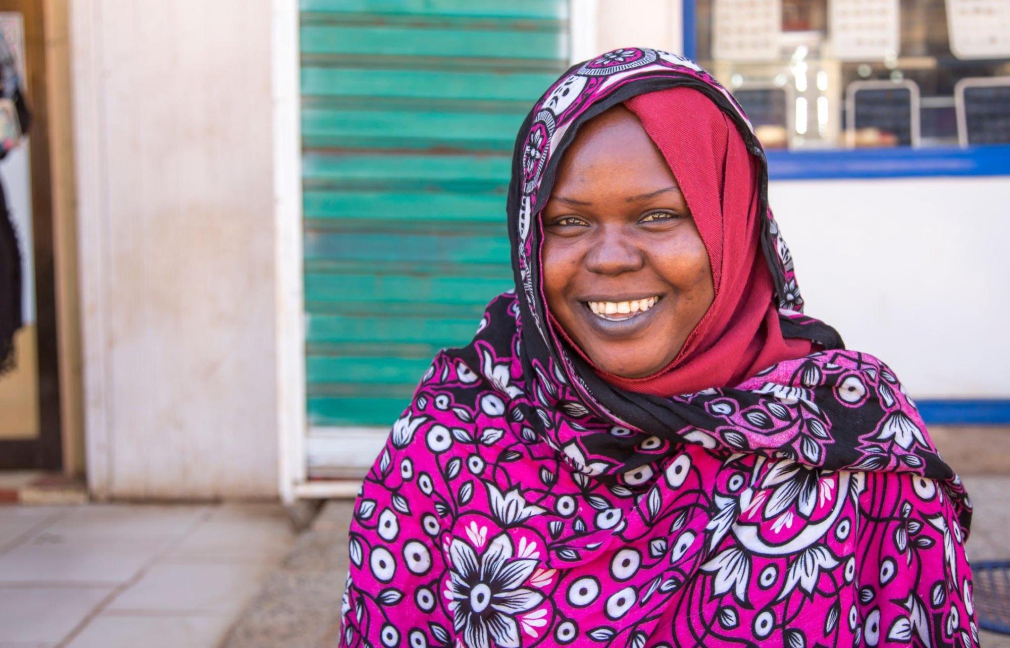 A smiling woman in a pink and black patterned headscarf sits outside a shopfront
